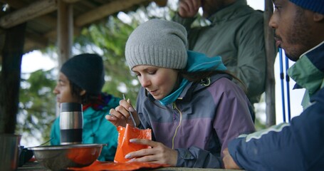 Adult African American man opens doy pack for his wife. Multiethnic tourist family eat hiking food together. Group of travelers or hikers rest in gazebo after expedition or trek in the mountains.