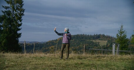 Caucasian woman stands on top of hill, takes selfie or shows beautiful nature view via video call using phone. Female tourist or hiker on vacation trip or hiking in the mountains. Outdoor recreation.