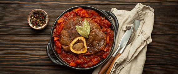 Traditional Italian dish Ossobuco all Milanese made with cut veal shank meat with vegetable tomato sauce served in black casserole pan top view on rustic brown wooden background.