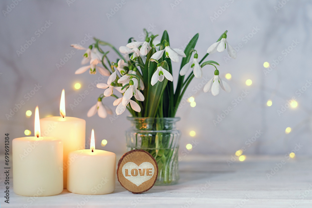 Wall mural bouquet of snowdrop flowers , heart decor and burning candles on table, abstract light background. s