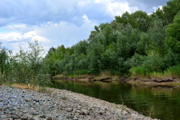 Fototapeta na wymiar riverbank with gravel and calm water and green trees before rain copy space