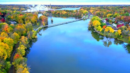 Eye catching Fall colors lining pristine blue river with walking path bridge, aerial view.
