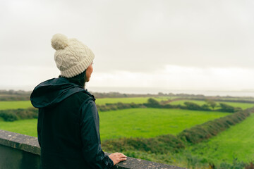 Unknown woman with a woolen hat observing a beautiful cloudy Irish landscape from a railing