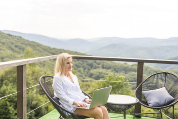 A young woman, freelancer is working on a laptop remotely on a balcony in the open fresh air near the mountains in warm sunny weather.