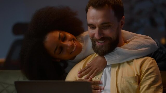 Focused bearded man working laptop while loving african american woman embracing him