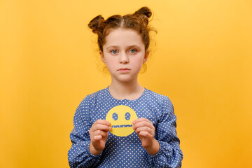 Portrait of unhappy upset preteen caucasian girl child holding sad yellow emoticon, looking at camera, posing isolated over plain yellow color background wall in studio. Feedback rating concept - obrazy, fototapety, plakaty