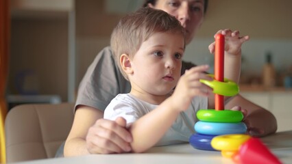 Obraz premium baby baby with mom collects a toy pyramid. happy family kindergarten kid dream concept. mom helps baby son collect a toy pyramid. son learns to lifestyle assemble a toy pyramid in the kindergarten