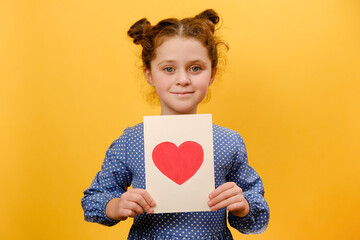 Portrait of pretty positive preteen girl child holding greeting card with heart symbol, happy looking at camera, posing isolated over yellow color background wall in studio. Father's Day concept