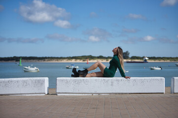 Young, beautiful, blonde woman, eyes closed and wearing a green dress, sitting on a stone bench, solitary, relaxed and calm, with the sea in the background. Concept of solitude, peace, tranquility.