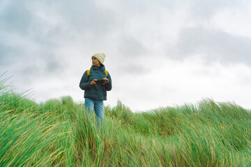 Woman traveling through Ireland with mobile phone in hand and winter hat among tall grass enjoying...