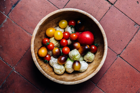Bamboo bowl full of fresh red and yellow cherry tomatoes and tomatillos, on the red and brown tiled floor
