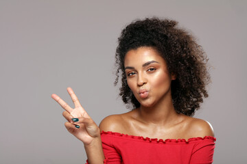 Playful positive curly haired woman send a kiss, shows a happy peace gesture.