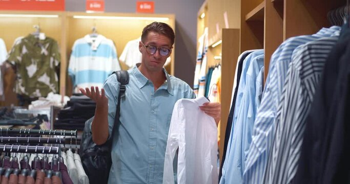 Man in men's clothing store closely examining shirt options Choice of shirt becomes challenging with high price tags. Surprise at cost during shirt selection moment of quality versus value.