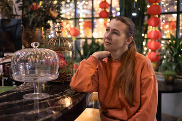 Middle-aged woman wearing peach fuzz coloured clothes sits in cafe decorated for Christmas and Chinese New Year. Chinese lanterns, glowing garlands. Festive mood. Self-love, self-care. Mental health