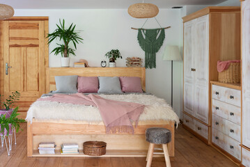 Boho interior of bedroom with wooden furniture and bed with pink blanket and pillows and plants. Wooden wardrobe in the cottage. Comfortable home.	