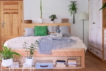 Boho interior of bedroom with wooden furniture and bed with grey pillows and green plants. ...