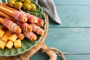 Slices of prosciutto or jamon. Delicious grissini sticks with prosciutto, cheese, rosemary, olives on green plate on dark background. Appetizers table with italian antipasto snacks Top view copy space