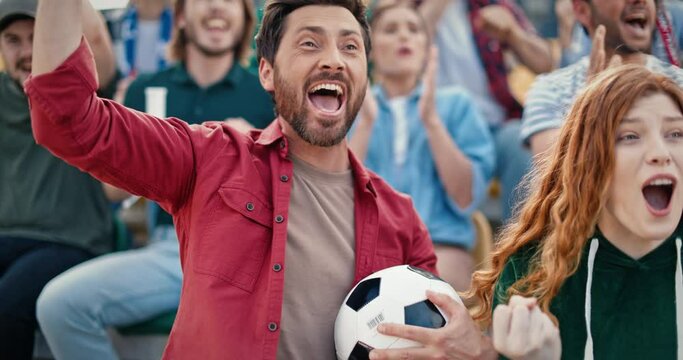 Beautiful Caucasian couple cheering for winners at football game. Handsome bearded man holding ball while sitting in stands. Victory celebration. People hugging each other and shouting joyfully.
