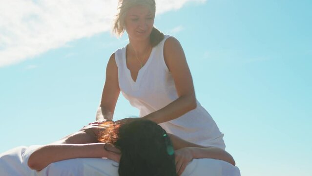 Low angle massage outdoor on special table. Caucasian female masseuse massages back, shoulder blades and neck of a dark-haired tanned woman with blue sky background. Healthy lifestyle and body care