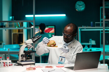 African american scientist examining hamburger with lab-grown meat sitting at his workplace in lab