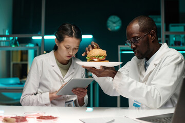 Black scientist examining hamburger with lab-grown meat, his colleague working on tablet nearby - obrazy, fototapety, plakaty