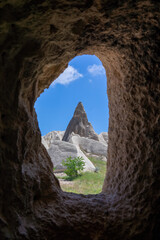 A view from within a cave, framing a lone fairy chimney against the vibrant blue sky and fluffy...