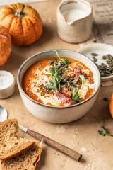 Bowl of pumpkin cream soup decorated with cream, pumpkin seeds, bacon, microgreens and olive oil on brown textured background with ingredients above