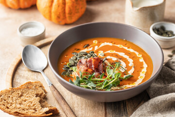 Pumpkin cream soup decorated with cream, pumpkin seeds, bacon, microgreens and olive oil on brown textured background with ingredients