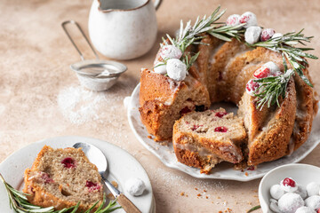 Christmas bundt cake decorated with glaze, cranberries and rosemary served on beige table. Homemade cake recipe