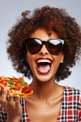 Happy woman with her mouth wide open in a smile, wearing stylish sunglasses. With curly hair and an infectious expression, she holds a delicious pan pizza in her hand