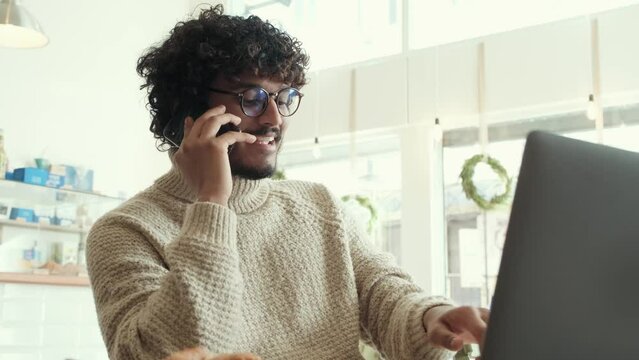 Cheerful curly haired indian man in beige sweater working on laptop and talking on cellphone in cafe