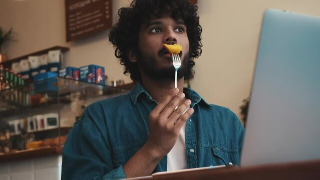 Pensive curly haired indian man wearing jeans shirt working on laptop and eating sponge cake in cafe