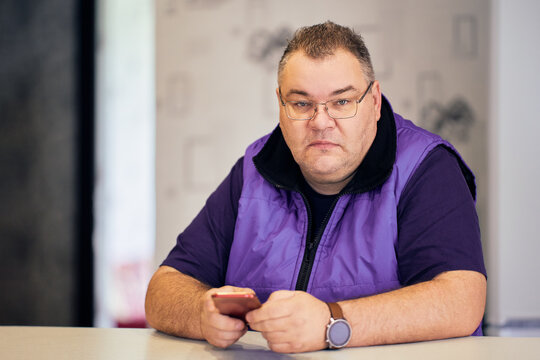 Plump serious man in his 40s wearing glasses and sleeveless jacket sits at table with phone in his hands.