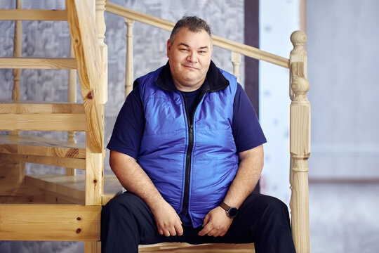 Sly smile on face of large European man, 47 years old, sitting on steps of stairs leading to second floor of his house.