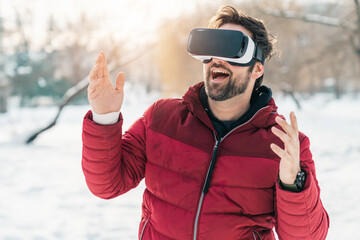 Excited man wearing virtual reality glasses gesturing with hands having fun outdoors on snowy day...