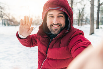 Happy smiling man waving hand holding the camera and taking a selfie outdoors wearing warm jacket standing in the snow.