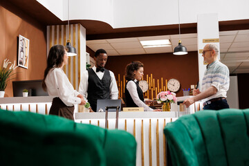Multiethnic employees welcome elderly guests at hotel reception with efficient service. Retired senior people checking in at counter. Hotel lobby providing comfortable atmosphere for visitors.