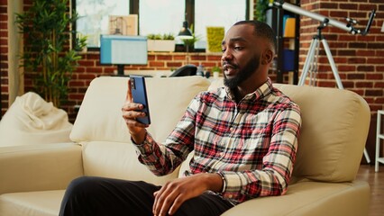 African american man enjoying day off at home, chatting with friends in video call. Smiling cheerful person excited to catch up with mates during online meeting while relaxing in cozy homely apartment