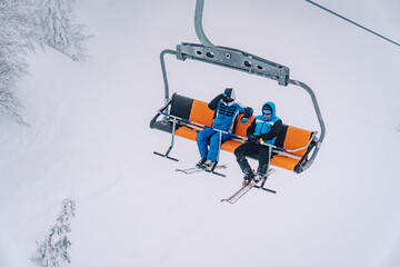 Skiers in ski suits and goggles ride on a chairlift above the snowy trees