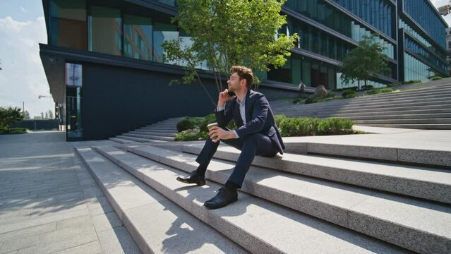 Businessman calling coffee break sitting stairs near office building zoom out.