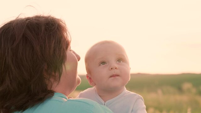 Happy family, mom, baby in nature. Mother kiss little son outdoor at sunset. Small kid in mothers arm look up at sky, smiles. Baby is sitt arms of parents on walk on summer day. Concept of motherhood