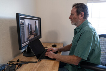 Mature Caucasian man sitting at a desk of a home office working on a computer with photo...