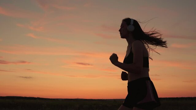 Silhouette, beautiful girl in musical headphones, is engaged in fitness, runs in evening. Training run. Free young woman runs in summer in park at sunset, listens to music on headphones. Teen runner