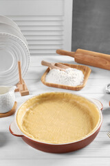 Pie tin with fresh dough, rolling pin and ingredients on white wooden table. Making quiche