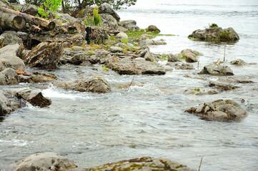 Stones and trunks of fallen trees on the shore of a large lake with a whirlpool surrounded by mountains.
