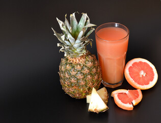 A mixture of fruit juices in a tall glass on a black background, next to pieces of ripe grapefruit and pineapple.