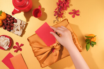 View from above of a hand stuffing a red envelope into a gift bag. A tea set, red orchids,...