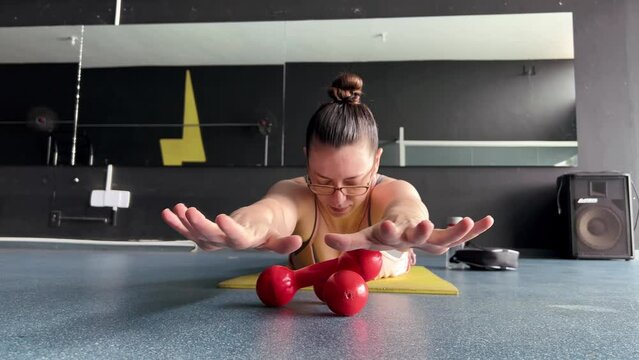 A woman lying face down on the mat in the gym, exercising with her hands in the air, a woman with glasses doing sports