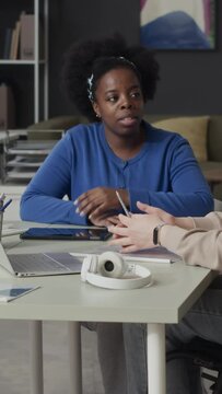 Vertical medium shot of young Black woman listening to Caucasian female colleague while sitting together at table in modern office