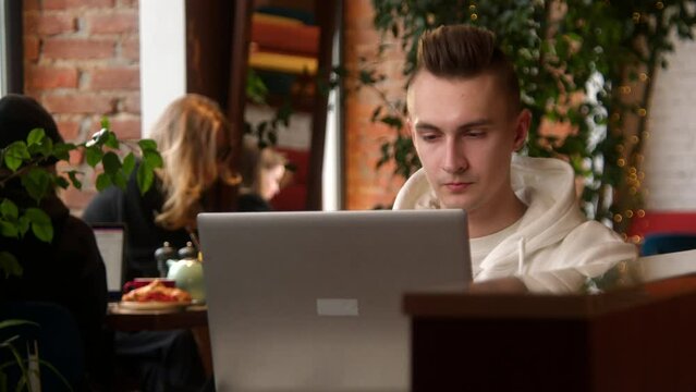 Young man is concentrating on laptop in cafe. Stock footage. Student is writing paper on laptop in cafe. Handsome young man freelancing at laptop in cafe